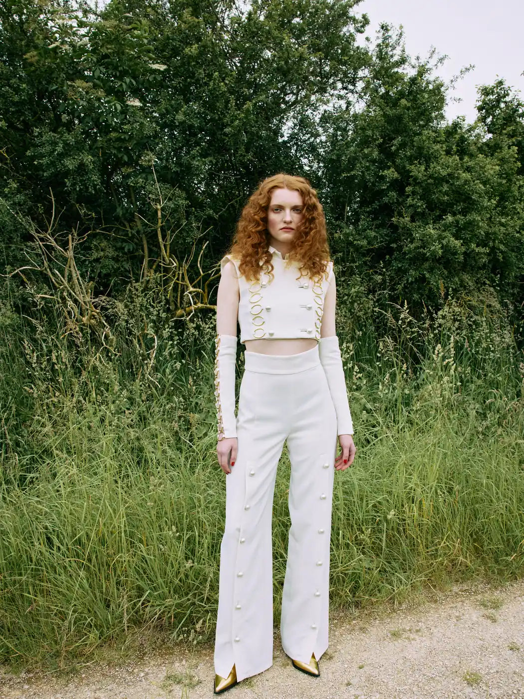 Model trägt Damen-Hose in Weiss mit hohem Bund, gerades Bein und durchgehende Knopfleiste am Bein mit weissen Perlenknöpfen. Dazu passende Weste und Armstulpen, Vorderansicht.