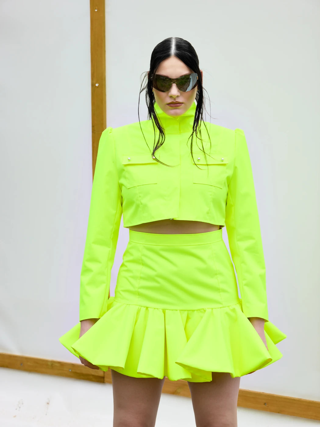 Model trägt Damen-Jacke in Neongelb und in cropped Länge mit hohem Stehkragen, Reißverschluss und aufgesetzten Brusttaschen mit Perlenknöpfen. Dazu passender Rock, Vorderansicht.