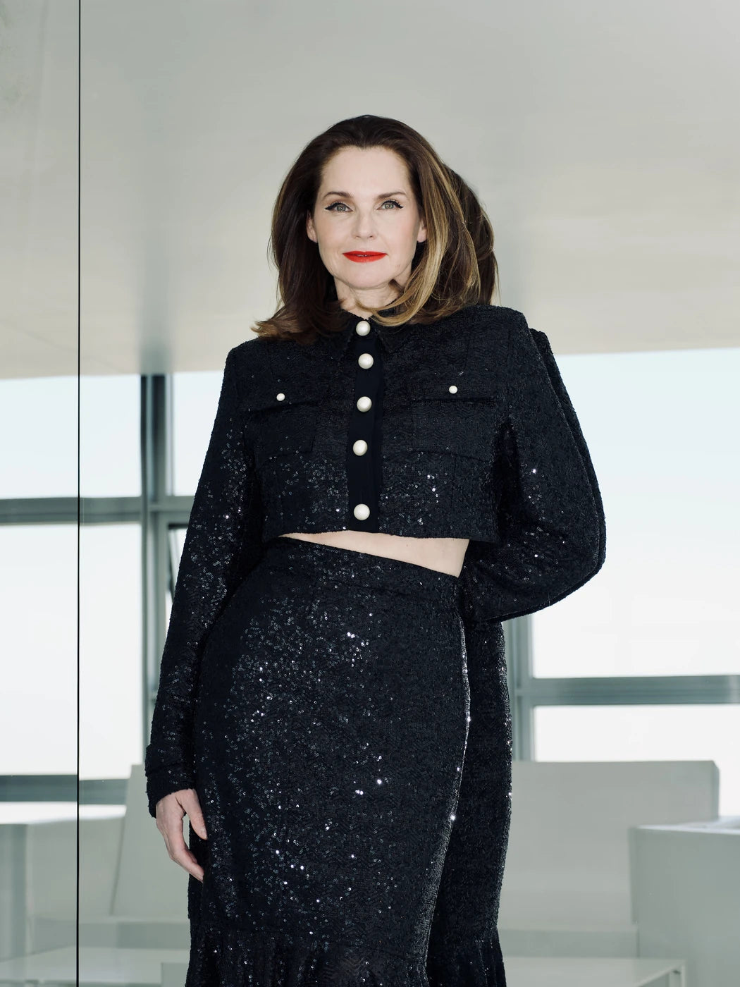 Model trägt Damen-Jacke mit Pailletten in Schwarz und in cropped Länge. Hemdkragen, Knopfleiste und aufgesetzte Brusttaschen mit Perlenknöpfen in Weiss. Dazu passender Rock, Vorderansicht.