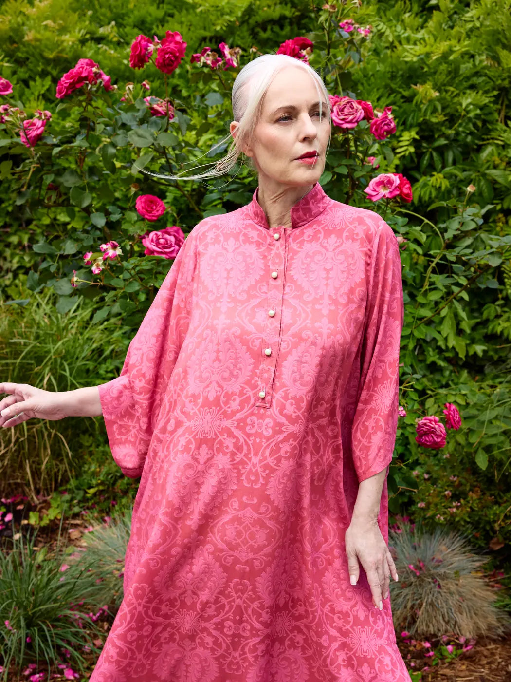 Model trägt Damen-Kaftankleid mit Ornament-Print in Korallenpink und Rosa, Stehkragen und Perlenknöpfen, Vorderansicht.