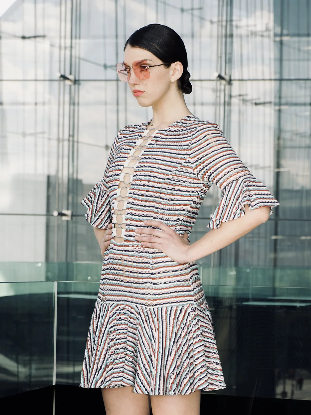 Model trägt Damen-Strickkleid mit Loch- und bunten Streifenmuster in Weiss, Türkis, Rosa, Braun und Schwarz, kurze Volantärmel und ausgestellter kurzer Saum, dazu Ringdetails auf Brusthöhe, Seitenansicht.