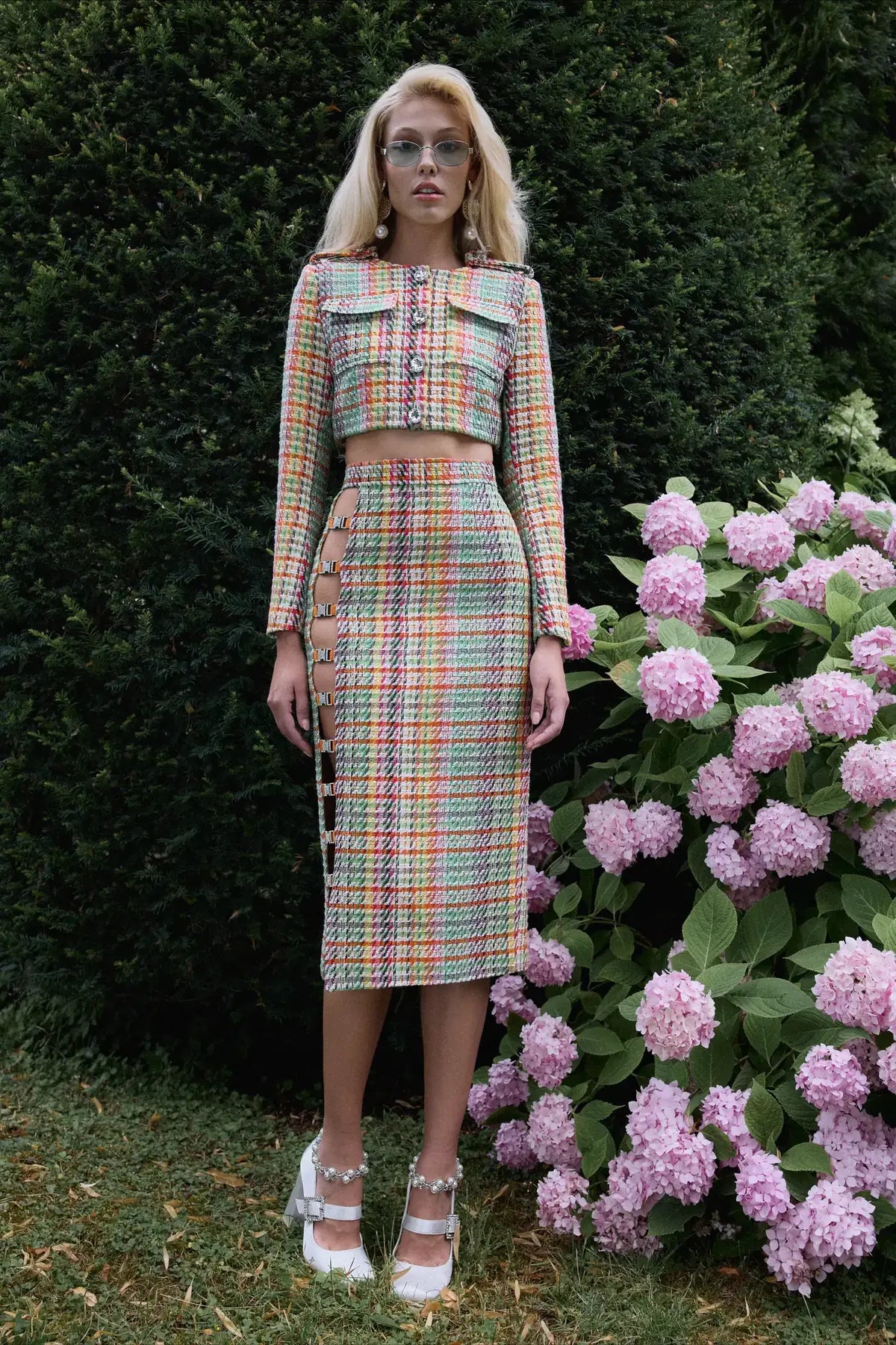 Kampagne Dancing Flowers: Damen-Set aus kurzer Tweedjacke und Midirock mit Schnallen, vor rosa Hortensien.