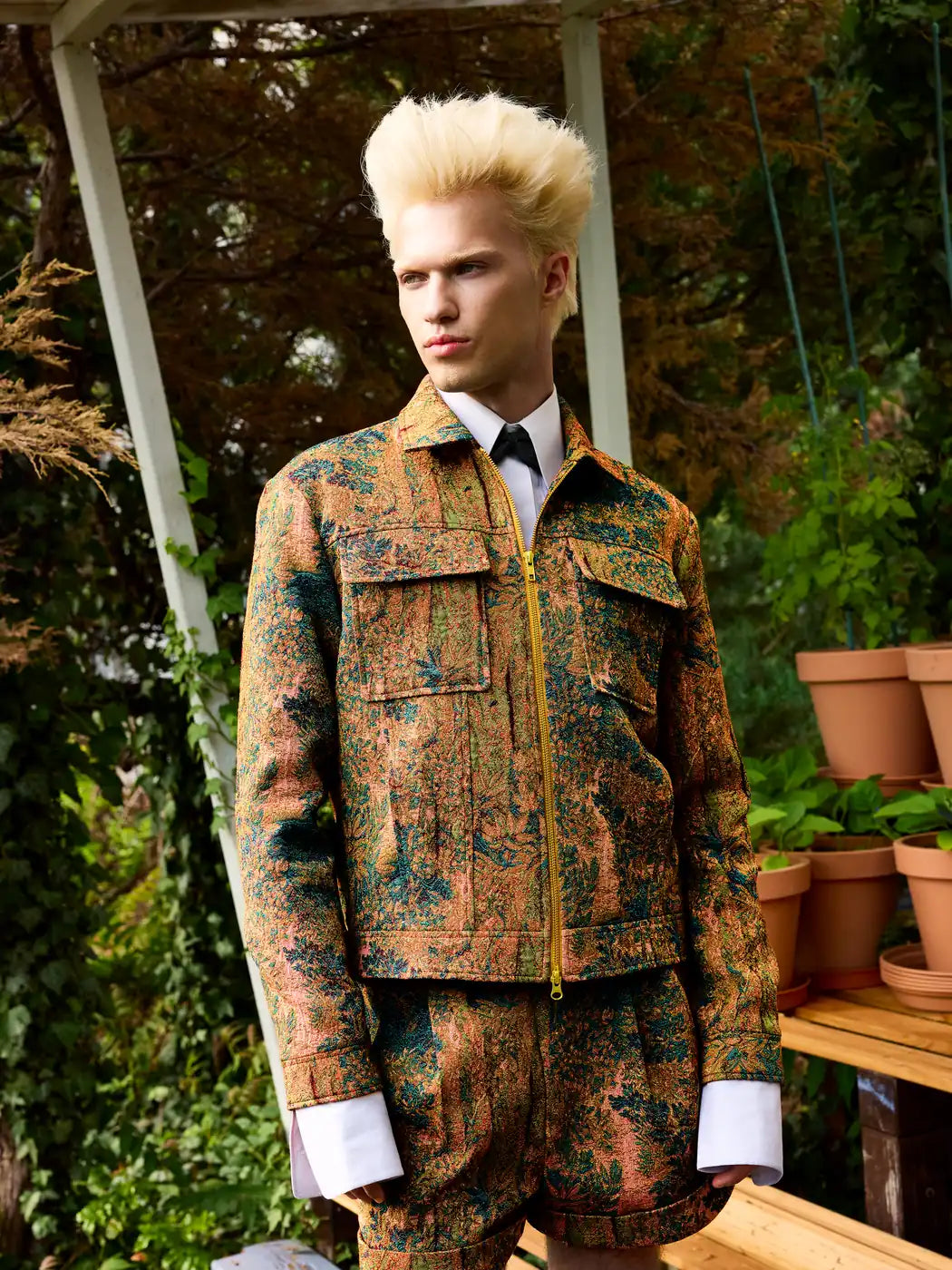 Model trägt Herren-Jacke aus Jacquard mit floralem Wald-Muster in Grün- und Kupfertönen, durchgehendem Goldreißverschluss und aufgesetzten Brusttaschen, Vorderansicht.